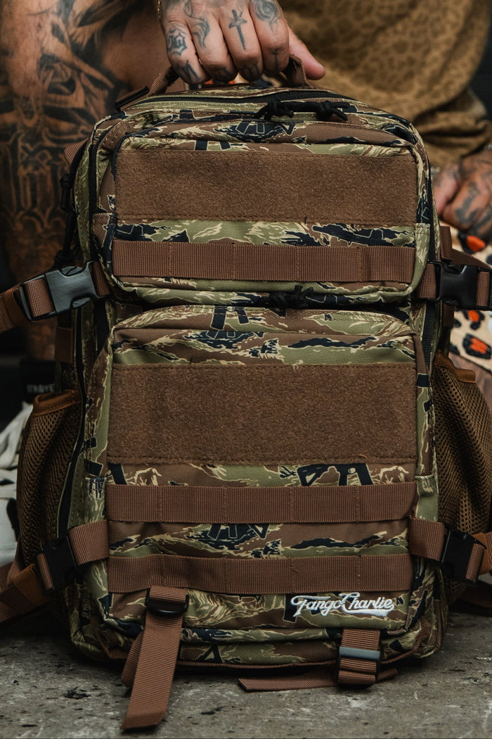 A tattooed individual sits outdoors on concrete, holding the Tango Charlie Apparel Snipes & Stripes - Tac Pac 25L backpack featuring brown straps and mesh pockets.