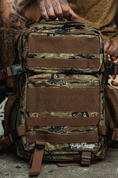 A tattooed individual sits outdoors on concrete, holding the Tango Charlie Apparel Snipes & Stripes - Tac Pac 25L backpack featuring brown straps and mesh pockets.
