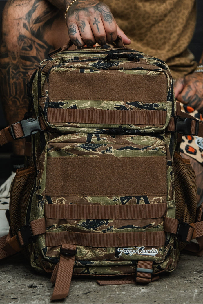 A person with tattooed hands holds the Snipes & Stripes - Tac Pac 25L, a camouflage backpack with brown straps and mesh pockets. The Tango Charlie Apparel logo is visible on the durable pack, which sits on a concrete floor.