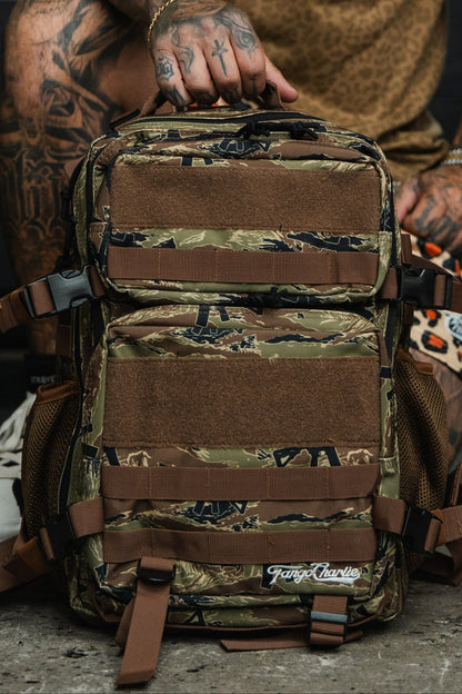 A person with tattooed hands holds the Snipes & Stripes - Tac Pac 25L, a camouflage backpack with brown straps and mesh pockets. The Tango Charlie Apparel logo is visible on the durable pack, which sits on a concrete floor.