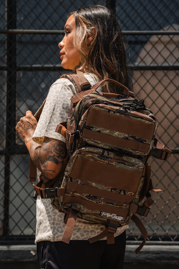 A person with long hair and tattoos, wearing a patterned t-shirt, stands outside by a metal fence carrying the Tango Charlie Apparel Snipes & Stripes - Tac Pac 25L backpack.