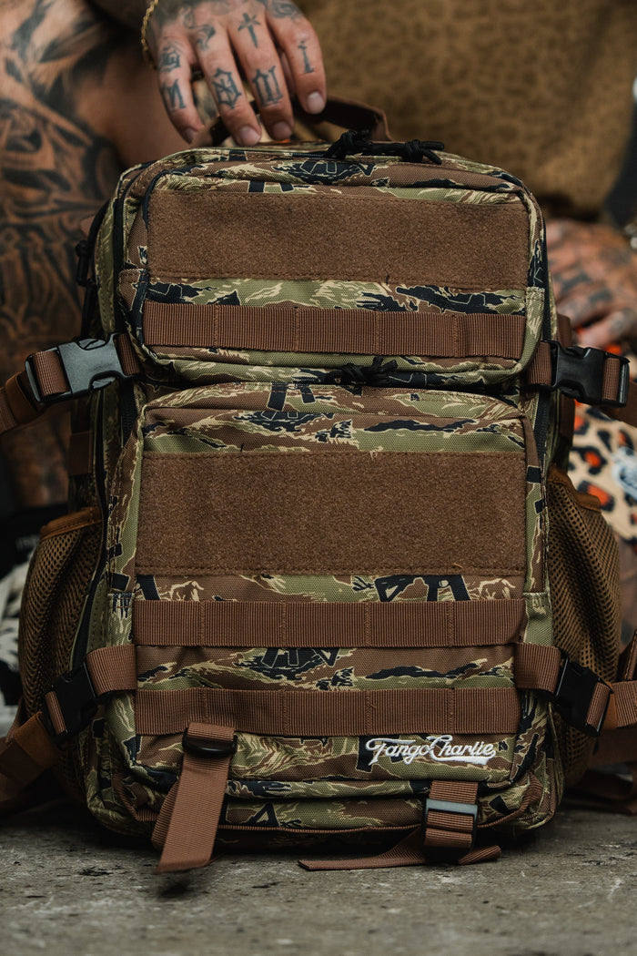 A tattooed person holds the Snipes & Stripes - Tac Pac 25L by Tango Charlie Apparel, a camouflage tactical backpack with brown straps and multiple compartments, designed for durability and organization, against a blurred background.