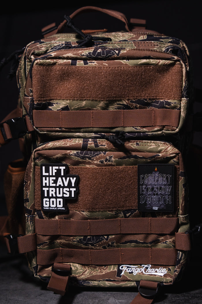 The Snipes & Stripes - Tac Pac 25L by Tango Charlie Apparel is a tactical backpack with brown straps, a camouflage design, and bold patches: "LIFT HEAVY TRUST GOD" in white and "COMFORT IS SLOW DEATH" in gothic text.