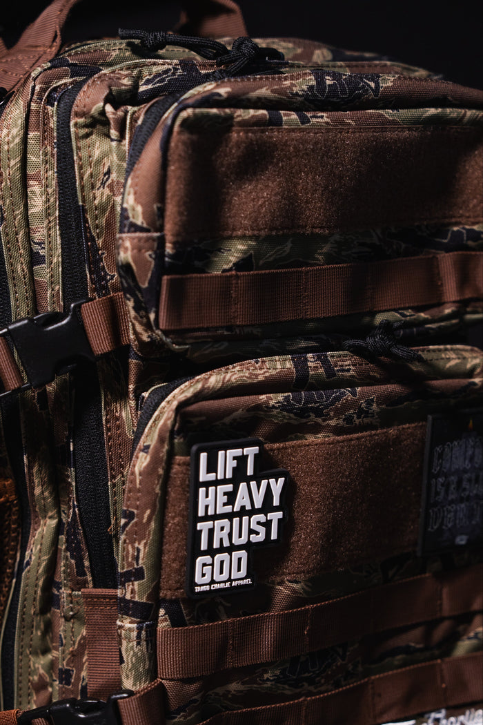 A close-up of the Snipes & Stripes - Tac Pac 25L by Tango Charlie Apparel shows a camouflage tactical backpack with brown straps. A front patch reads “LIFT HEAVY TRUST GOD” in bold white letters. The dark background highlights its rugged style.