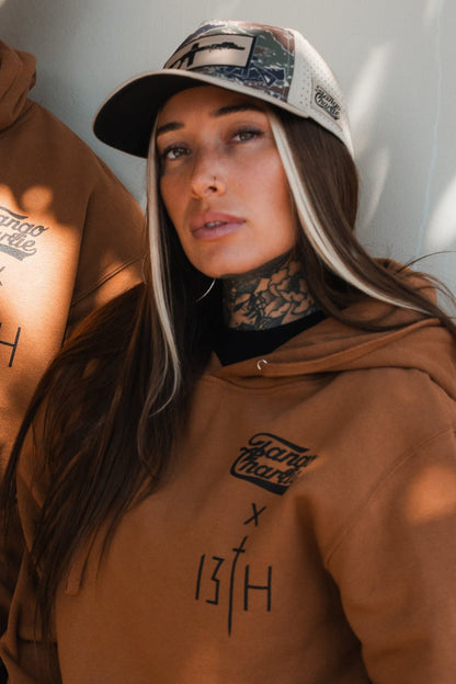 A woman with long dark hair and light streaks, wearing a brown hoodie and the Snipes & Stripes Hydro Snapback Hat by Tango Charlie Apparel, poses against a light background, showing tattoos on her neck.