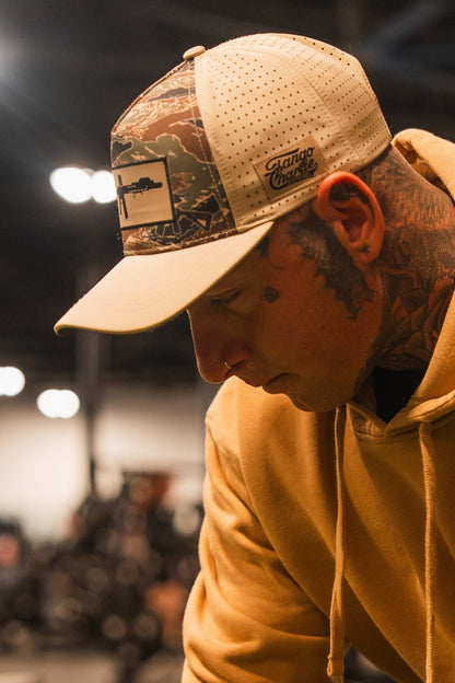A man with face and neck tattoos looks down in soft indoor light, wearing a tan hoodie and the Snipes & Stripes Hydro Snapback Hat by Tango Charlie Apparel. The blurred background features lights and indistinct shapes.