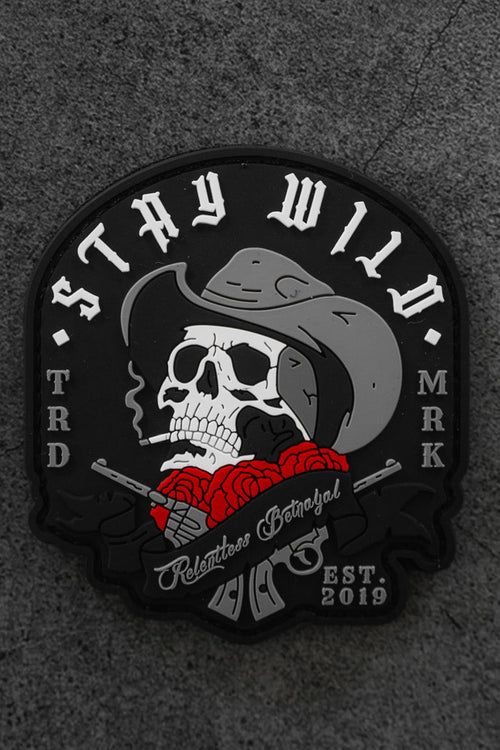 The Stay Wild Patch by Relentless Betrayal is a black Velcro patch featuring a skull in a cowboy hat smoking a cigarette, red roses, crossed revolvers, and the text “STAY WILD,” “Relentless Betrayal,” and “EST 2019.”.