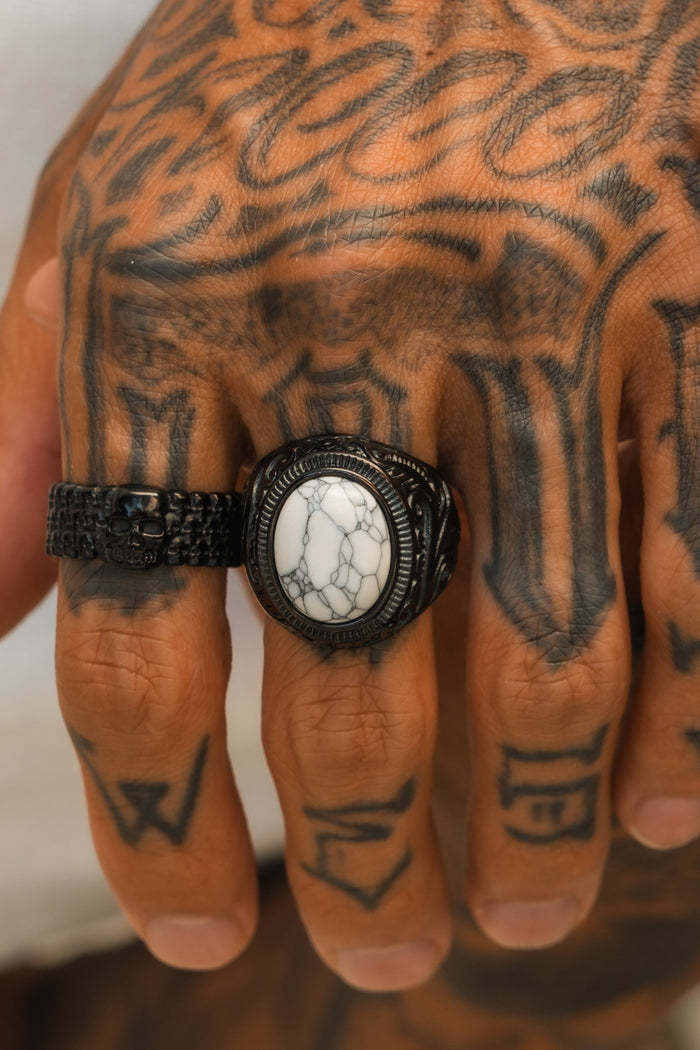 A tattooed hand wearing two black rings, including the Relentless Betrayal Storm Cloud Ring—handcrafted stainless steel with a marble-like stone and textured skull—set against a neutral background.