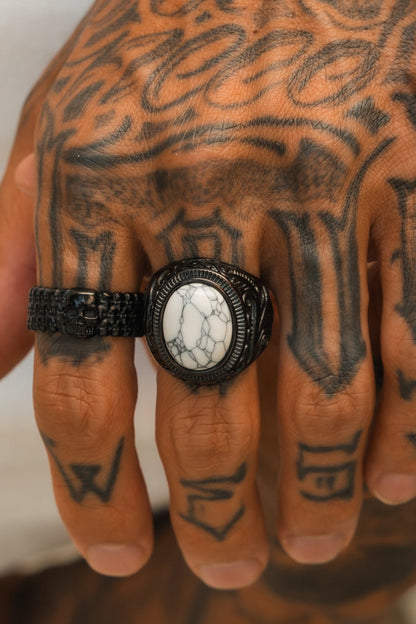 A tattooed hand wearing two black rings, including the Relentless Betrayal Storm Cloud Ring—handcrafted stainless steel with a marble-like stone and textured skull—set against a neutral background.