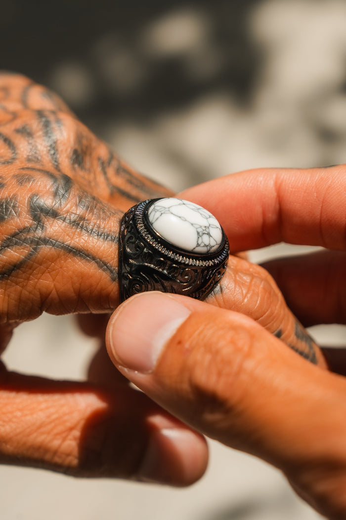 Tattooed hands hold the Storm Cloud Ring by Relentless Betrayal, a handcrafted stainless steel piece with an intricate design and a large white gemstone streaked with gray veins.