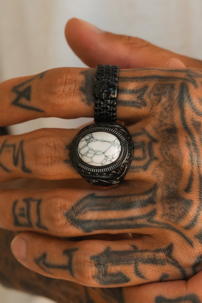 A hand with detailed black tattoos wears the Relentless Betrayal Storm Cloud Ring, a bold handcrafted stainless steel piece set with a large round white stone veined in dark, held by an ornate textured band.