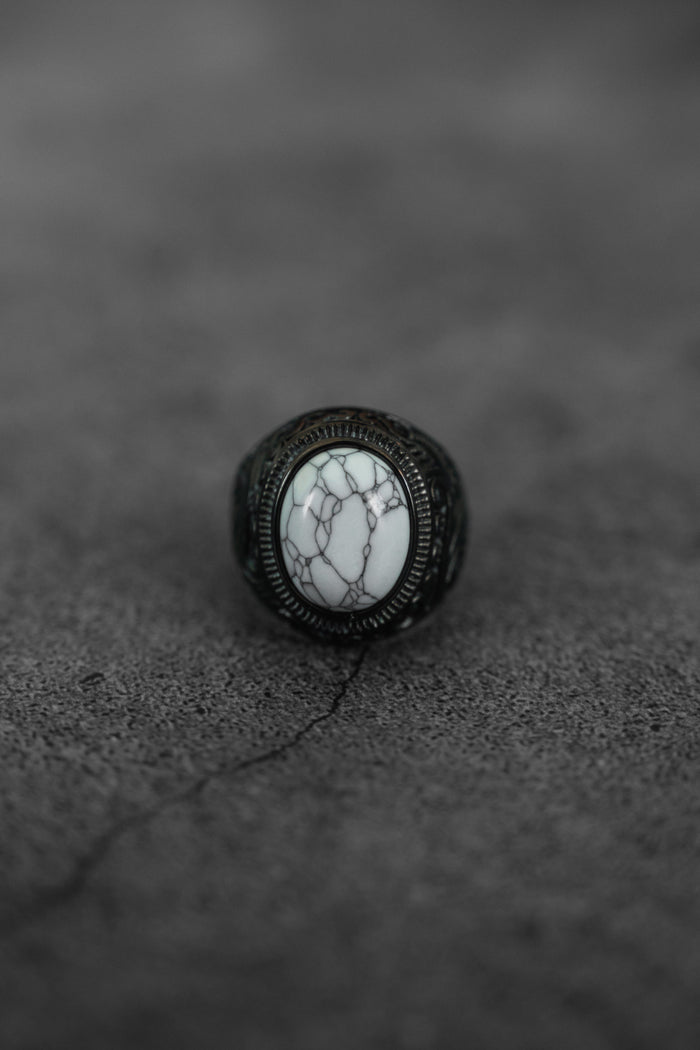 The Storm Cloud Ring by Relentless Betrayal features a large oval white stone with dark gray veining, set in an ornate blackened metal band, showcased on a cracked gray textured surface.