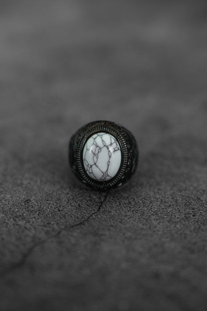 The Storm Cloud Ring by Relentless Betrayal features a large oval white stone with dark gray veining, set in an ornate blackened metal band, showcased on a cracked gray textured surface.
