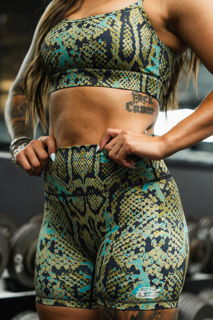 A woman at the gym wears the Tango Charlie Apparel Straight Strap Sports Bra | Lethal Venom in green and yellow snakeskin print, paired with matching shorts, revealing toned abs and a side tattoo, with dumbbells blurred in the background.