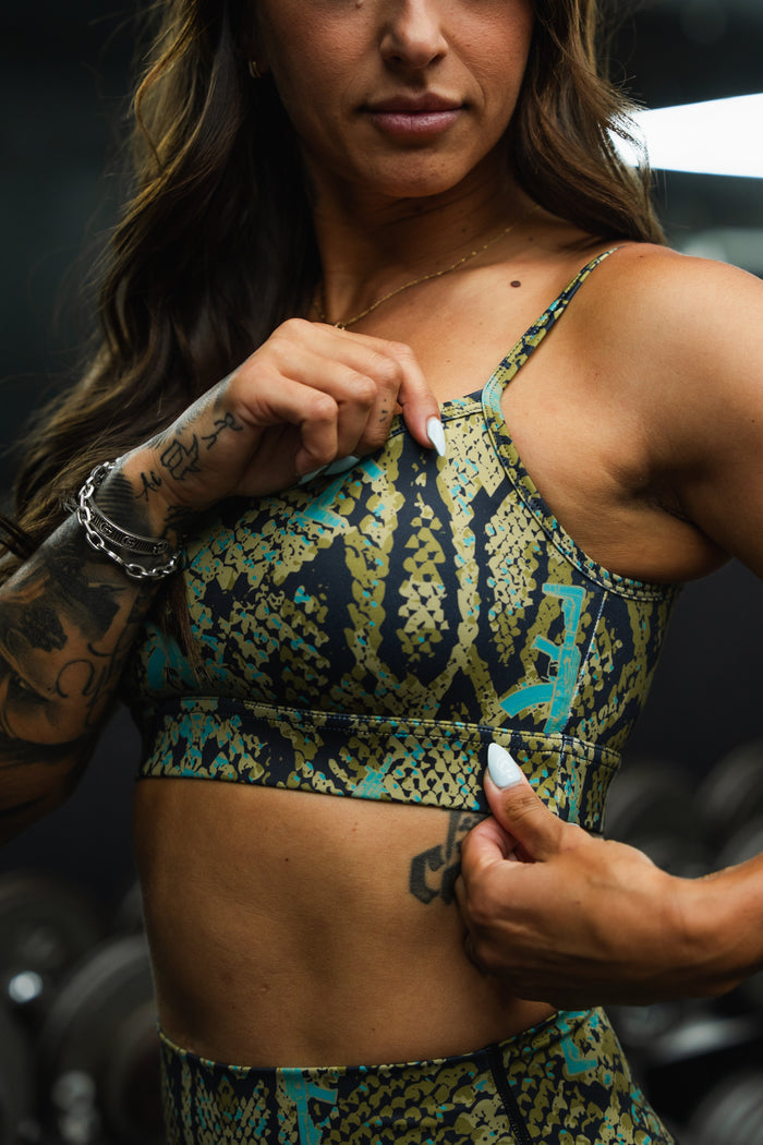A woman in the Tango Charlie Apparel Straight Strap Sports Bra | Lethal Venom, featuring a green and black pattern, adjustable straps, and moisture-wicking fabric, adjusts her top to reveal tattoos, with blurred gym equipment in the background.