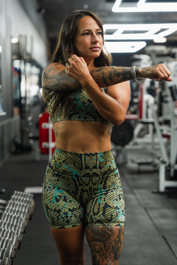 A woman in the Tango Charlie Apparel Straight Strap Sports Bra | Lethal Venom stretches her arm across her chest in a gym, surrounded by fitness equipment and dumbbells.