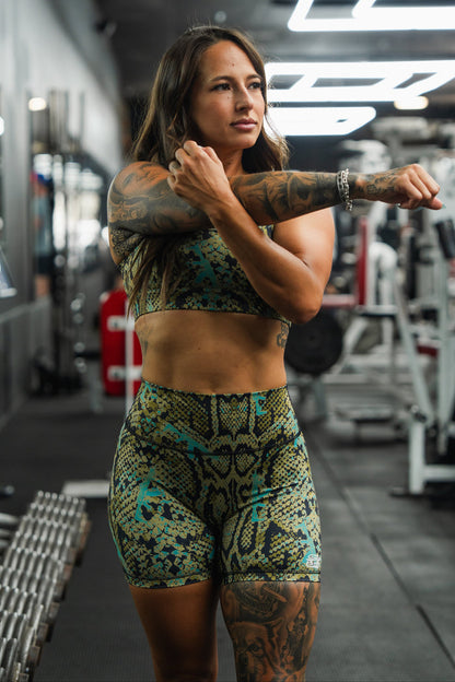 A woman in the Tango Charlie Apparel Straight Strap Sports Bra | Lethal Venom stretches her arm across her chest in a gym, surrounded by fitness equipment and dumbbells.