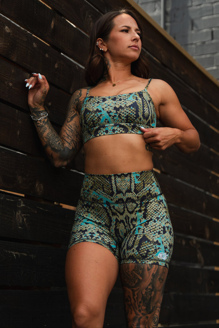 A tattooed woman poses confidently against a dark wooden wall, wearing the Straight Strap Sports Bra | Lethal Venom by Tango Charlie Apparel—a medium support, moisture-wicking set in green, blue, and yellow tones.