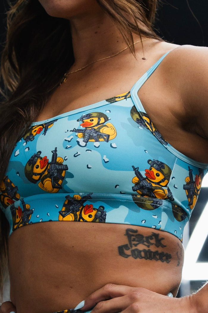 A woman models the Born United Private Label Straight Strap Sports Bra in TactiQuack, featuring adjustable straps and a cartoon duck soldier print. Her ribcage tattoo reading "F@ck Cancer" is visible; only her upper body appears.