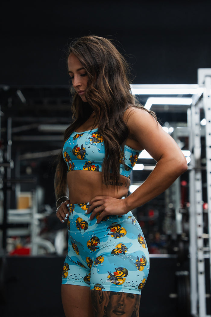 A woman with long brown hair stands in a gym wearing Born United Private Label’s Women's High-Waisted Seamless Front Compression Training Shorts by TactiQuack, featuring blue fabric with cartoon bee patterns, as she looks down with hands on hips.