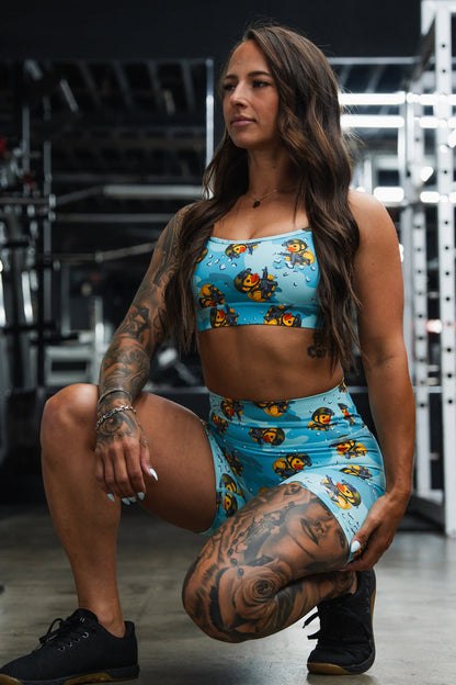A woman with long brown hair and tattoos kneels confidently in a gym, wearing Born United Private Label's Women's High-Waisted Seamless Front Compression Training Shorts by TactiQuack. Gym equipment appears in the background.