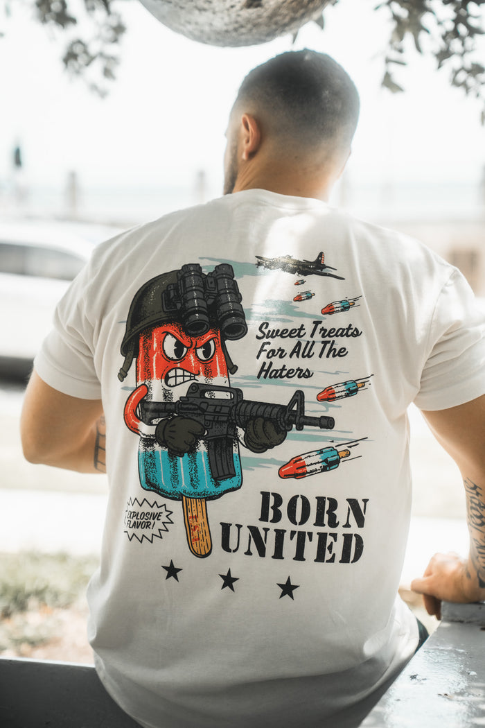 A man sits outdoors with his back to the camera, wearing a Born United Private Label "Sweet Victory" T-shirt featuring a cartoon popsicle with night vision goggles and fighter jets, with the text “Sweet Treats For All The Haters” and “BORN UNITED.”.