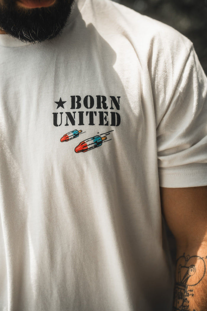 A person wearing the "Sweet Victory" white t-shirt by Born United Private Label, featuring BORN UNITED text and two small rockets on the chest. The image shows part of their beard and a tattooed arm.