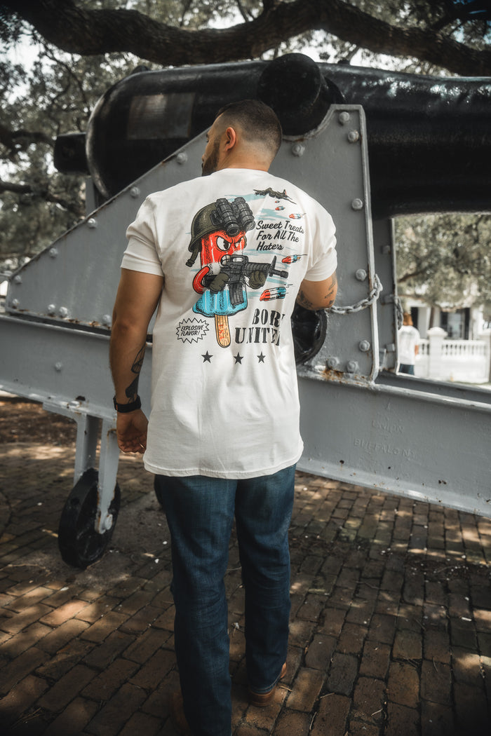 A man outdoors in jeans and a white Born United Private Label “Sweet Victory” T-shirt with a colorful popsicle graphic and text on the back stands before a large cannon, with trees and brick pavement behind him.