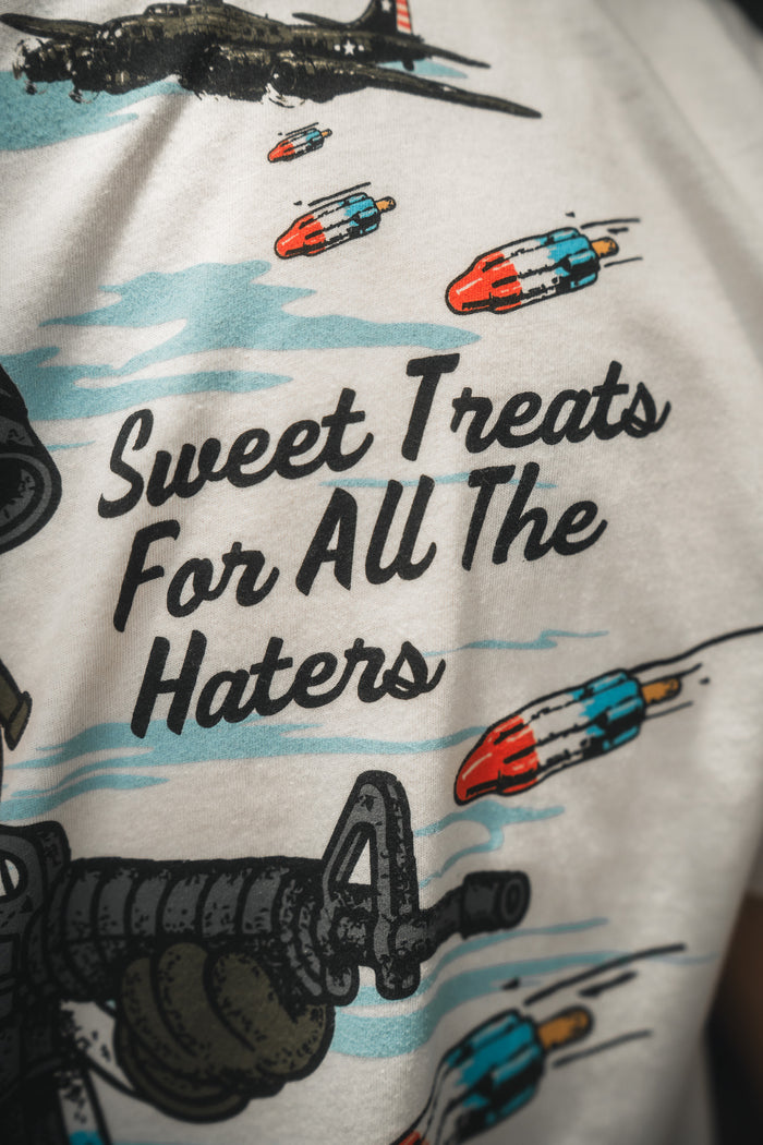 The Sweet Victory T-shirt by Born United Private Label is white, features colorful cartoon rockets, bold “Sweet Treats For All The Haters” text, and includes part of an airplane in the design.