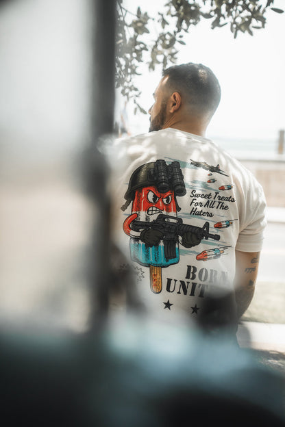A man with short hair and a beard is seen from behind, wearing a Sweet Victory T-shirt by Born United Private Label featuring a cartoon ice pop with a gun, helmet, and the text Sweet Treats For All The Haters.