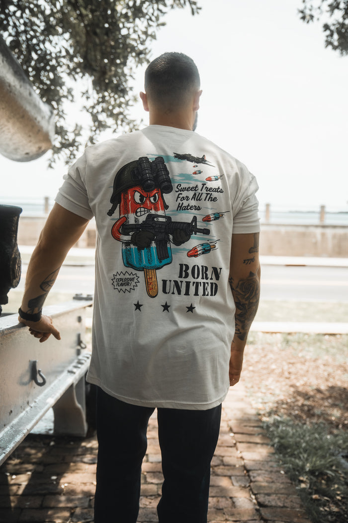 A man stands outdoors facing away, wearing the Born United Private Label "Sweet Victory" white T-shirt with a cartoon popsicle, rockets, and “Sweet Treats For All The Haters” text. Trees and a street are visible in the background.