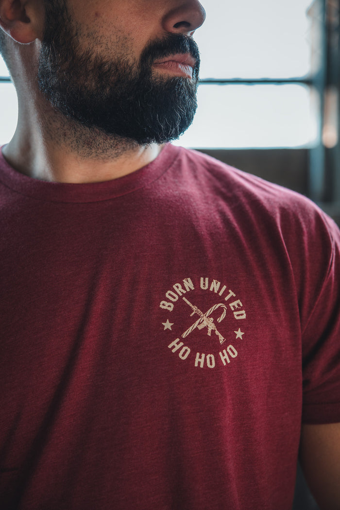 A bearded man sports the Born United Private Label Tactical Santa Tee, a maroon limited-edition shirt with "BORN UNITED HO HO HO" and a rifle graphic on the chest, giving off an armed Saint Nick vibe.