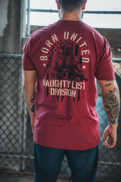 A person in a limited-edition Born United Private Label Tactical Santa Tee, featuring an armed Saint Nick and "NAUGHTY LIST DIVISION" on the back, stands near a metal fence.