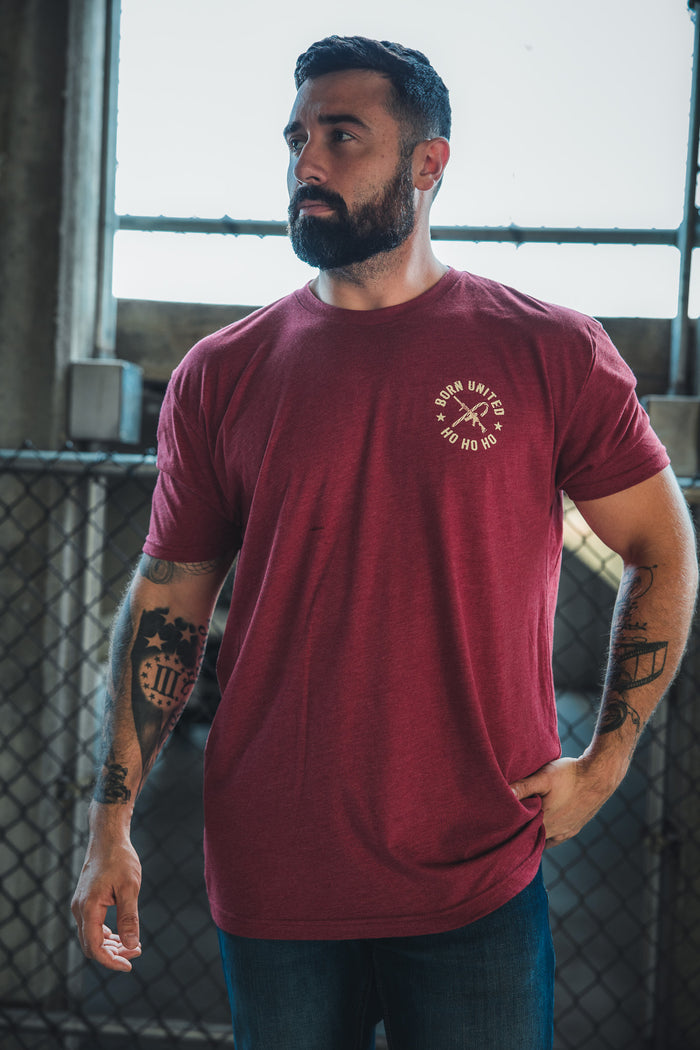 A bearded man wears the Born United Private Label Tactical Santa Tee, a limited-edition red Christmas shirt featuring a small printed design, while standing by a chain-link fence and window in an industrial setting.