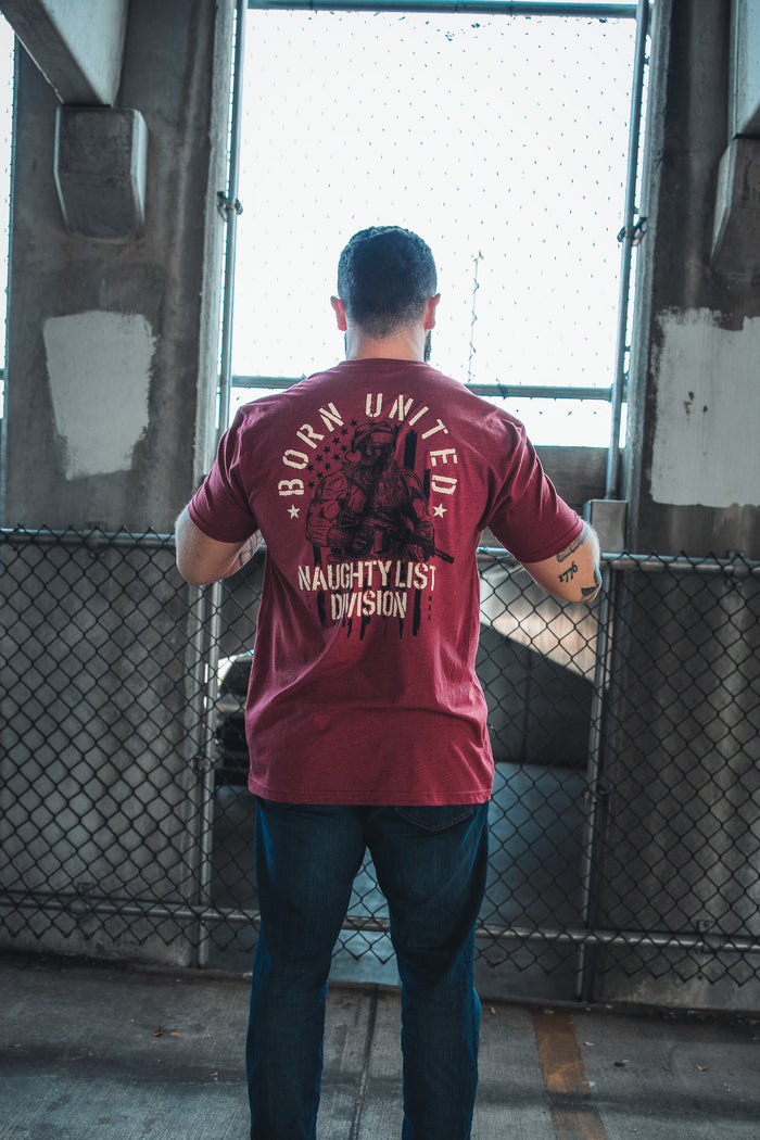 A person stands facing away, wearing a limited-edition red Tactical Santa Tee from Born United Private Label, in front of a chain-link fence and concrete pillars.