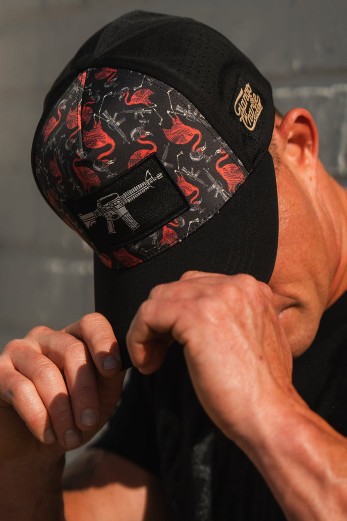 A person in a Tango Charlie Apparel Tactimingo Hydro snapback, a black cap with pink flamingos and a gun patch, tilts it down to obscure their face. The sweat-resistant hat adds flair as they stand against a concrete wall.