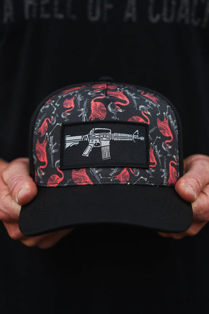 An individual holds a Tango Charlie Apparel Tactimingo Hydro snapback hat, featuring a sweat-resistant red and gray design with guns and buffalo, centered by a patch illustrating a white rifle.