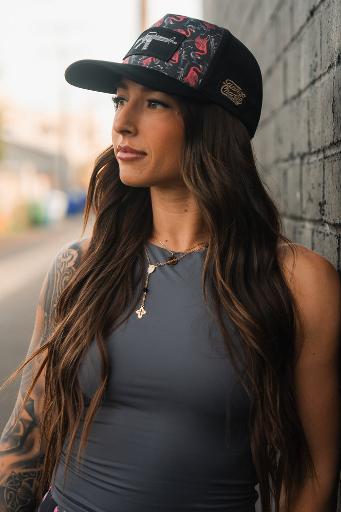 A woman with long brown hair wearing a Tango Charlie Apparel Tactimingo Hydro Snapback leans against a brick wall, displaying a tattoo on her right arm. She sports a sweat-resistant gray sleeveless top to stay cool, her serene and confident gaze directed to the side.