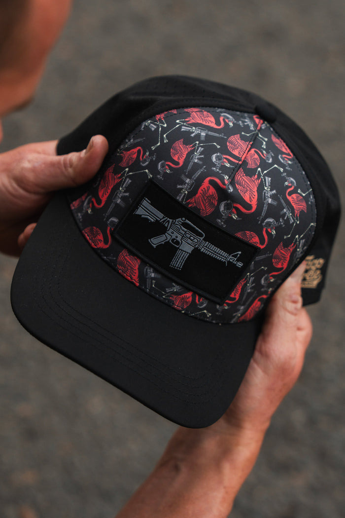 Hands holding the Tango Charlie Apparel Tactimingo Hydro Snapback Hat, showcasing a sweat-resistant black cap with a red flamingo and cactus pattern, featuring a rifle patch on the front against a softly blurred outdoor scene.