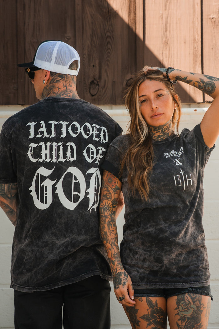 Two tattooed people wear matching Relentless Betrayal Tattooed Child of God Premium Mineral Wash T-Shirts—he shows the back print, she faces forward, both displaying ink in bold Christian style.