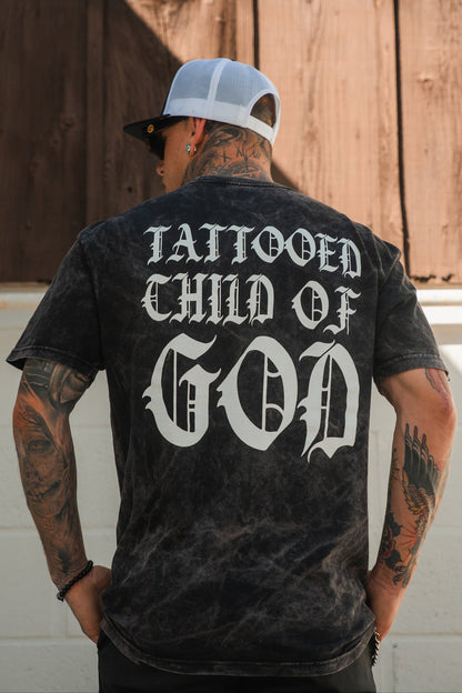 A tattooed individual wears the Relentless Betrayal Tattooed Child of God Premium Mineral Wash T-Shirt, featuring bold white gothic back lettering, paired with a white and black cap—a standout Christian apparel choice.