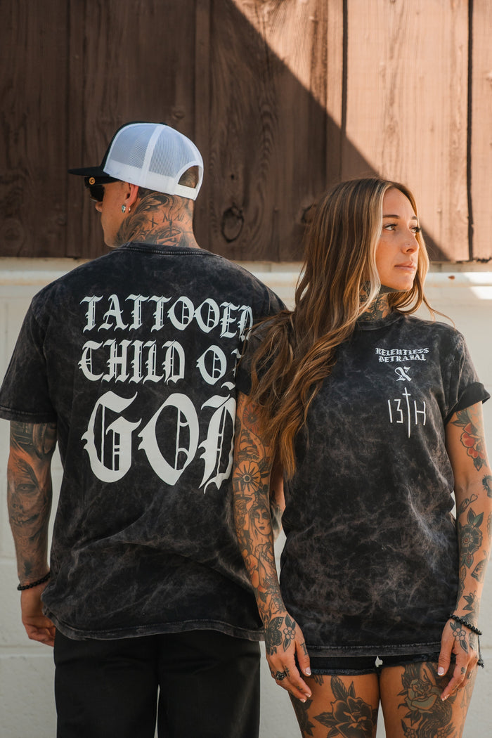 A tattooed man and woman in black marble-wash outfits stand side by side. He wears a Relentless Betrayal Tattooed Child of God Premium Mineral Wash T-Shirt; her top displays “Heights Steeple XIX 13TH.”.