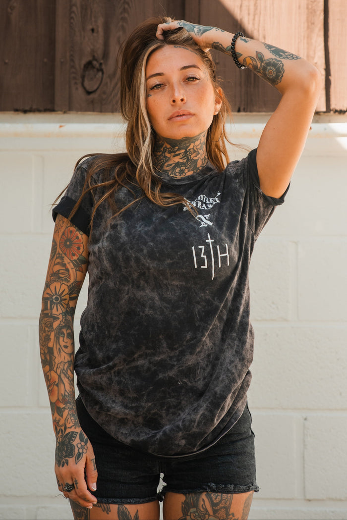 A woman with long hair and visible tattoos stands against a white brick wall, wearing the Relentless Betrayal Tattooed Child of God Premium Mineral Wash T-Shirt and black shorts, boldly showcasing Christian apparel.