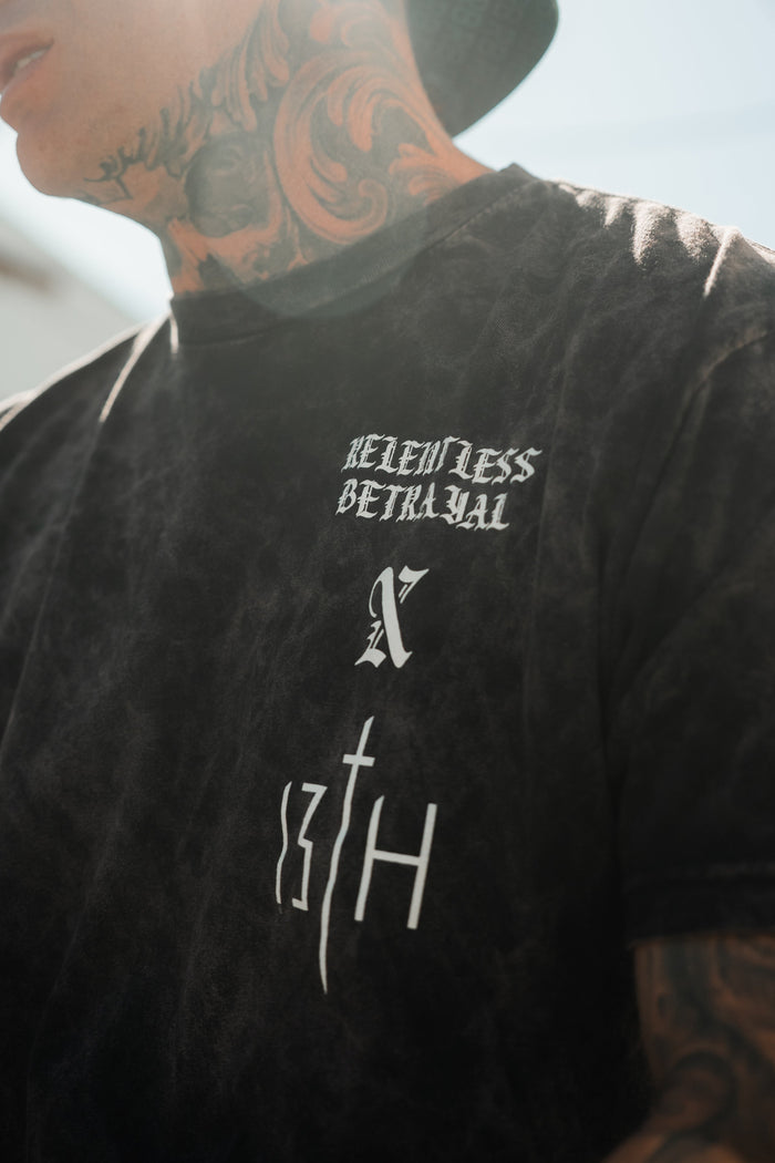 A tattooed Child of God wears the Relentless Betrayal Tattooed Child of God Premium Mineral Wash T-Shirt in black, with bold white text and a stylized X—Christian apparel for those who express faith with edge. Face not fully visible.