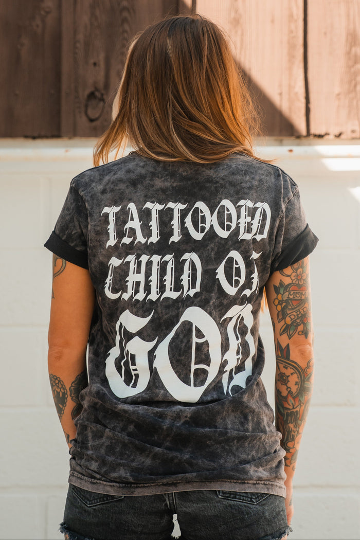 Seen from behind, a person with tattooed arms wears the Relentless Betrayal Tattooed Child of God Premium Mineral Wash T-Shirt, featuring bold white text and standing out as unique Christian apparel.