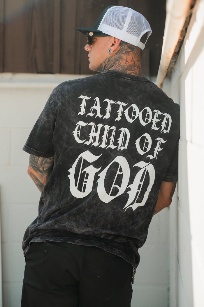 A tattooed, pierced man wears a Relentless Betrayal Tattooed Child of God Premium Mineral Wash T-Shirt with bold white text on the back, plus a mesh cap and sunglasses, standing against a white wall.