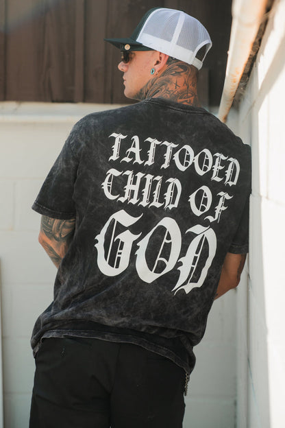 A tattooed, pierced man wears a Relentless Betrayal Tattooed Child of God Premium Mineral Wash T-Shirt with bold white text on the back, plus a mesh cap and sunglasses, standing against a white wall.