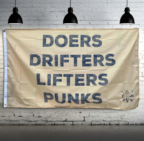 The Team Doers Drifters Lifter Punk Flag by Tango Charlie Apparel features bold text and hangs on a white brick wall. It includes metal grommets and double-stitching, illuminated by three black industrial-style lights above.