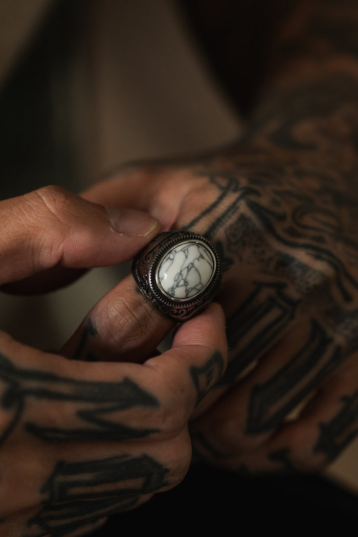 A person with tattooed hands, reminiscent of elemental forces, holds the Tempest Ring by Relentless Betrayal. This ring is steeped in mythical tales and features a marbled white stone set in a detailed dark metal band.