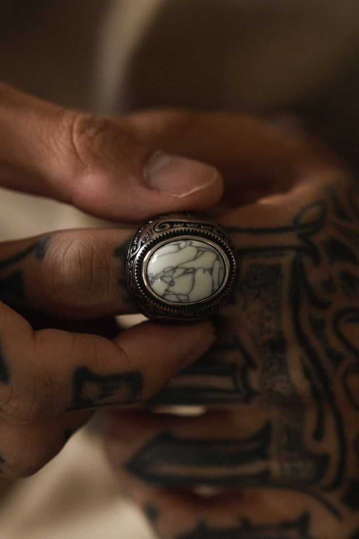 A close-up shows tattooed hands holding a Relentless Betrayal Tempest Ring. The ornate band cradles a large, smooth white stone veined with dark lines, subtly resting between thumb and forefinger, evoking elemental forces from mythical tales.